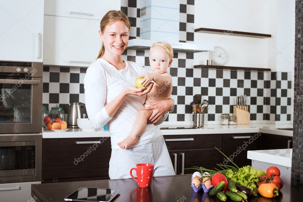 Mother Holding Daughter In Kitchen while cooking a meal of fresh vegetables and fruits. Mother and little daughter on kitchen holding an apple. Healthy food and healthy living concept