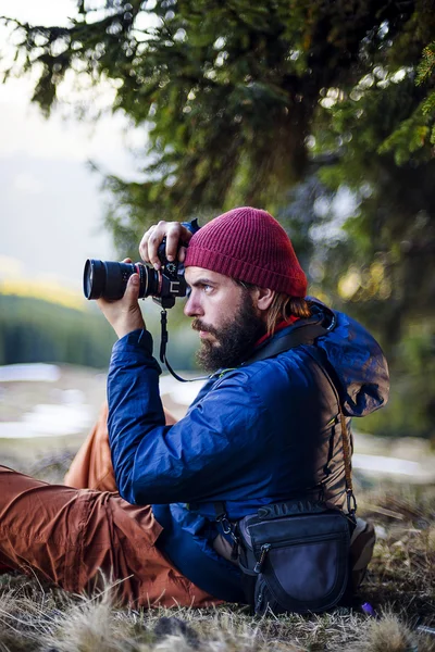 Młody brodaty mężczyzna z aparatu w drewno, fotograf robienia zdjęć w górach — Zdjęcie stockowe