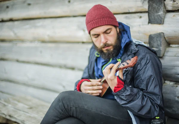 Brodaty facet grając ukulele w górach w pobliżu drewniany dom — Zdjęcie stockowe