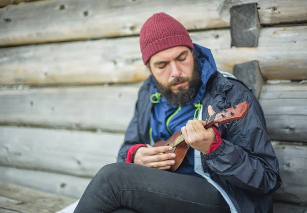 Brodaty facet grając ukulele w górach w pobliżu drewniany dom — Zdjęcie stockowe