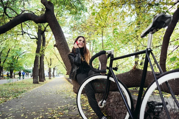 Joven hembra con bicicleta sentada en un árbol y hablando por teléfono — Foto de Stock