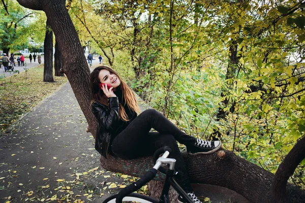 Jovem fêmea com bicicleta sentada em uma árvore e falando ao telefone — Fotografia de Stock