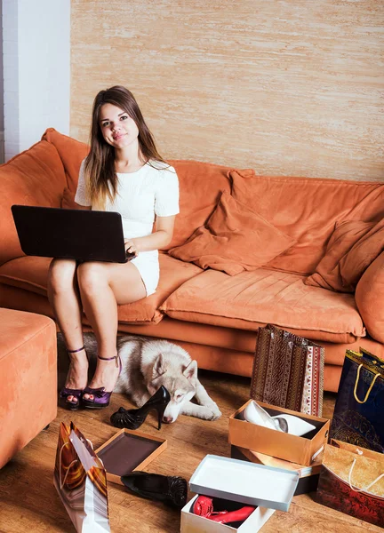 young caucasian female with laptop and shopping bags in living room. Woman tired of shopping. Female and little husky dog after shopping at home