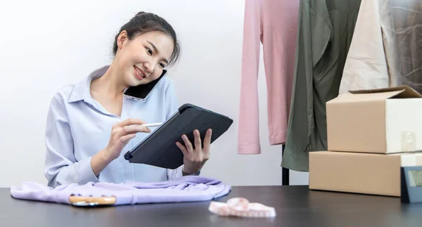 Genç Asyalı bir kadın akıllı telefon kullanarak müşteriyle sohbet ediyor ve siparişi onaylamak için tablet kullanıyor, çevrimiçi kıyafet satış işi, kıyafet satışı veya internet üzerinden satış işi.