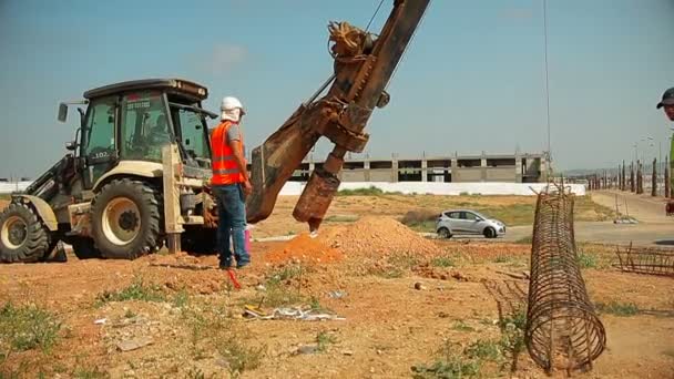 Τρακτέρ ανελκυστήρες μία χαλύβδινη ράβδο και το βάζουμε σε μια τρύπα στο έδαφος. Οι εργαζόμενοι στις κατασκευές βοηθώντας τον. — Αρχείο Βίντεο