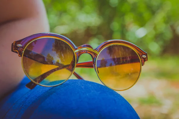棕色的太阳眼镜躺放在大腿上，一名穿蓝色牛仔裤的女子，坐在外面的草地上 — 图库照片