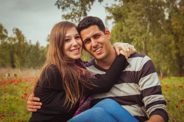 Pareja joven sentada en la hierba en un campo de amapolas rojas y — Foto de Stock