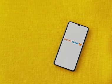 Lod, Israel - 8 Temmuz 2020: Meuhedet Sağlık Hizmetleri uygulaması sarı kumaş arka planda siyah bir cep telefonunun logosu İbranice metin ile çalıştırma ekranı. Üst görünüm düzlüğü kopyalama alanı ile yatıyordu.