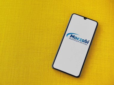 Lod, Israel - 8 Temmuz 2020: Maccbi Sağlık Hizmetleri uygulama ekranı üzerinde sarı kumaş arka planda siyah bir cep telefonunun logosu var. Üst görünüm düzlüğü kopyalama alanı ile yatıyordu.