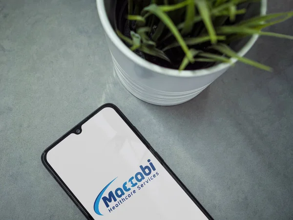 Lod, Israel - 8 Temmuz 2020: Siyah cep telefonlu modern minimalist ofis çalışma alanı, Maccbi Sağlık Hizmetleri uygulama ekranı ve mermer zemin üzerinde logosu var. Üst görünüm düzlüğü kopyalama alanı ile yatıyordu.