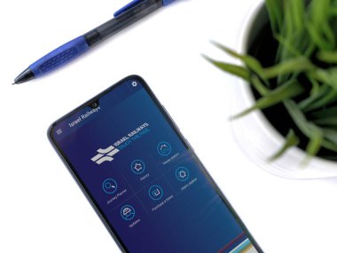 Lod, Israel - 8 Temmuz 2020: Siyah cep telefonlu modern minimalist ofis çalışma alanı ve beyaz arka planda logosu olan İsrail Demiryolları uygulaması fırlatma ekranı. Üst görünüm düzlüğü kopyalama alanı ile yatıyordu.