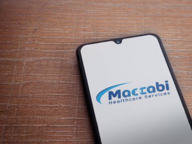 Lod, Israel - 8 Temmuz 2020: Maccbi Sağlık Hizmetleri uygulama ekranı üzerinde siyah bir cep telefonunun ahşap arka planda sergilendiği logo. Üst görünüm düzlüğü kopyalama alanı ile yatıyordu.