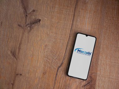 Lod, Israel - 8 Temmuz 2020: Maccbi Sağlık Hizmetleri uygulama ekranı üzerinde siyah bir cep telefonunun ahşap arka planda sergilendiği logo. Üst görünüm düzlüğü kopyalama alanı ile yatıyordu.