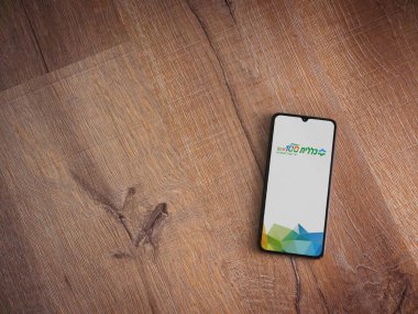 Lod, Israel - 8 Temmuz 2020: Clalit Sağlık Hizmetleri uygulama ekranı İbranice metinlerle siyah bir cep telefonunun ahşap arka planda sergilenmesi ile başlatıldı. Üst görünüm düzlüğü kopyalama alanı ile yatıyordu.