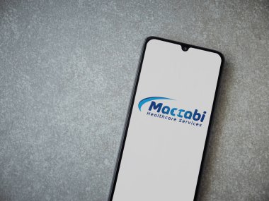 Lod, Israel - 8 Temmuz 2020: Maccbi Sağlık Hizmetleri uygulaması seramik taş zemin üzerinde siyah bir cep telefonunun logosu olan fırlatma ekranı. Üst görünüm düzlüğü kopyalama alanı ile yatıyordu.