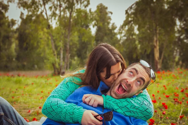 Junges Paar, das auf dem Gras in einem Feld aus roten Mohnblumen sitzt. das Mädchen küsst den Kerl, während er ein dummes Lächeln hat. — Stockfoto
