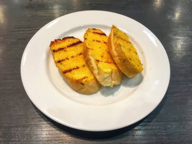 Tahta masadaki beyaz tabakta servis edilen üç dilim kızarmış sarımsaklı ekmek..