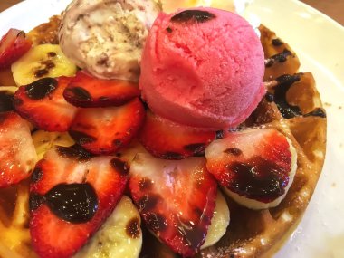 Dilimlenmiş çilek, muz, çikolata şurubu ve bir kepçe çilekli ve vanilyalı dondurmayla kaplı taze pişmiş waffle, bozulmuş bir tatlı yaratıyor..