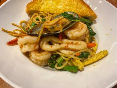 Baharatlı deniz ürünleri spagettisi. Karıştırılmış karides, kalamar, midye ve otlar. Beyaz tabakta bir dilim sarımsaklı ekmekle servis ediliyor..