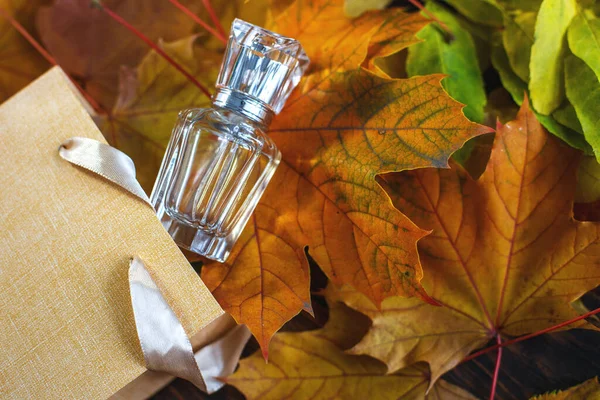 A glass bottle of female perfume with gift bag laid on autumn yellow leaves on a wooden background. Natural perfumery. Autumn season.