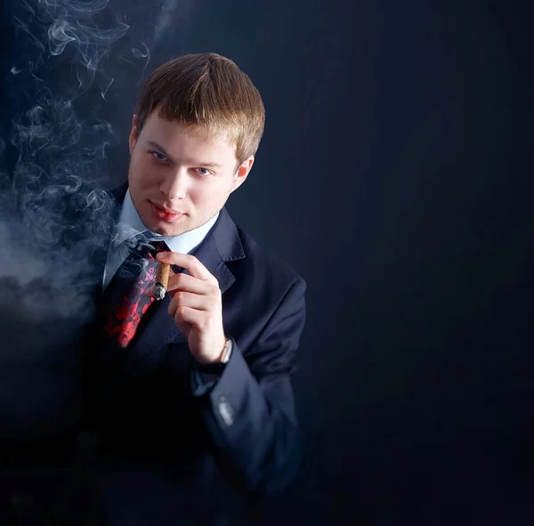 Ο άνθρωπος στο κοστούμι καπνίζουν ένα πούρο, μας κοιτάζει. τα μέρη του καπνού. Σκούρο φόντο — Φωτογραφία Αρχείου