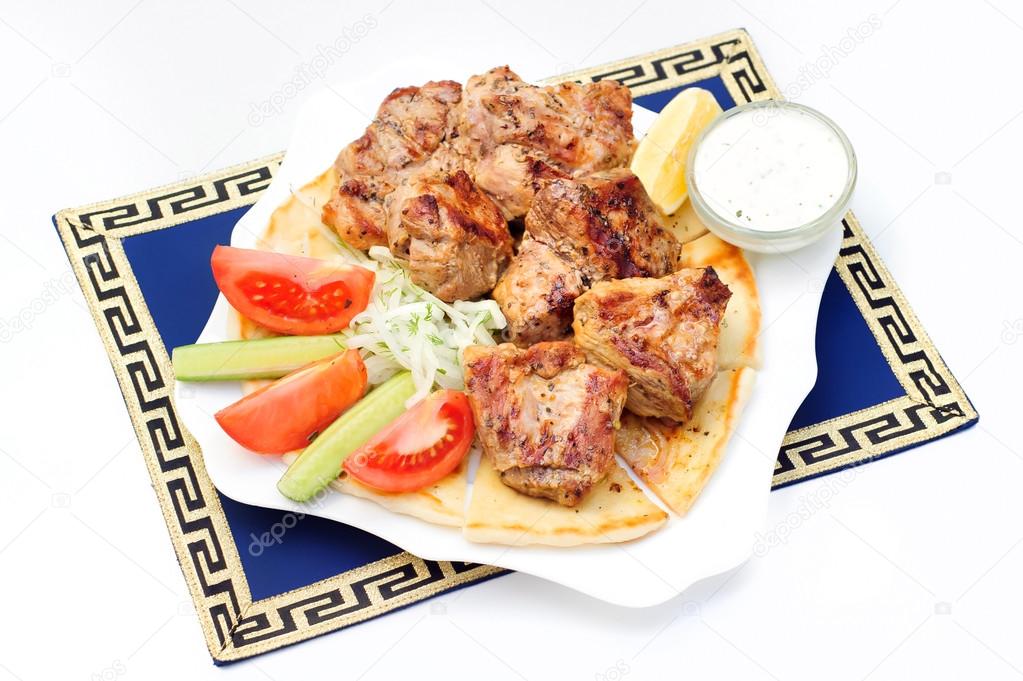 Souvlaki, kebab, grilled meat on pita bread with sauce, tomatoes and cucumbers, white plate.