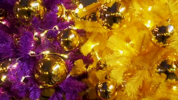 Волшебный красивый рождественский и новогодний фон. Искусственные цветные елки с большими золотыми шариками и мерцающими рождественскими огнями. Фиолетовый и желтый искусственные ели закрыть задний план — стоковое видео