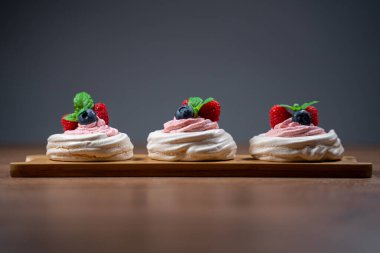 Üç mini Pavlova keki ve içi taze böğürtlen ve ahşap yüzeyde nane yapraklarıyla süslenmiş çilekli krema. Gri arkaplanda izole