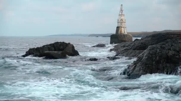 海浪冲击着深灰色的悬崖峭壁 拍打着浪花 后面是白色的灯塔和遥远的海岸线 多云的一天 保加利亚黑海沿岸的阿赫托波尔 — 图库视频影像