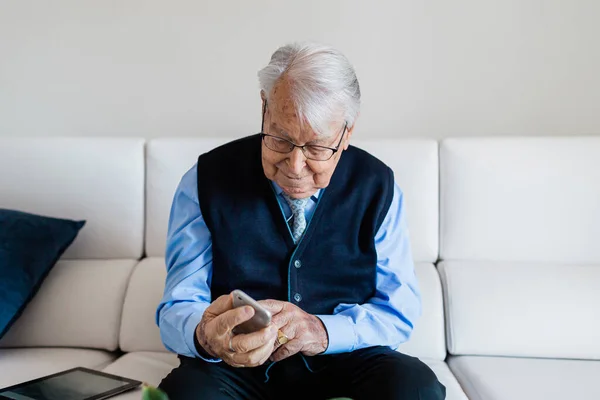 Elderly man dressed in blue shirt and tie, finishing talking on cell phone sitting on white leather sofa