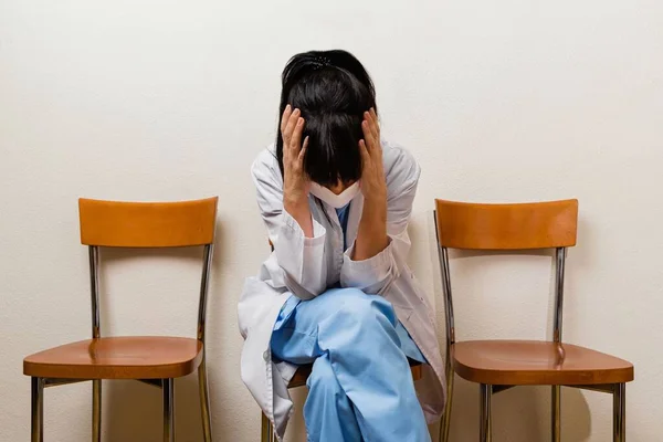 A very sad and tired nurse holding her head with two hands, sitting in a waiting room after her shift at the hospital.