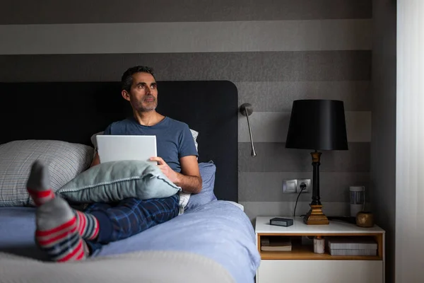Man in pajamas lying on bed reading with a digital tablet
