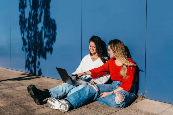 two long-haired friends, one blonde and one brunette, sitting on the floor in front of a blue wall working on a laptop.