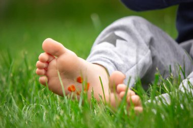 Çocuk çimlerin üzerinde oturuyor, boyalarla çocuğun bacağına gülümsüyor.