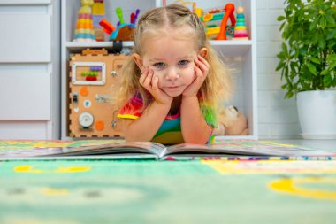 Parlak elbiseli küçük bir kız evde renkli bir paspasın üzerinde oyun oynuyor ve öğreniyor.