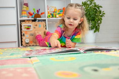 Parlak elbiseli küçük bir kız evde renkli bir paspasın üzerinde oyun oynuyor ve öğreniyor.