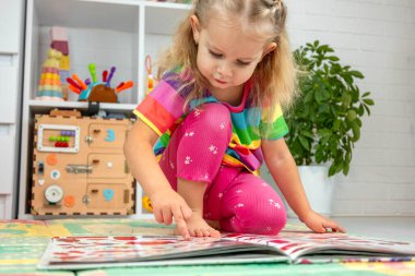 Parlak elbiseli küçük bir kız evde renkli bir paspasın üzerinde oyun oynuyor ve öğreniyor.