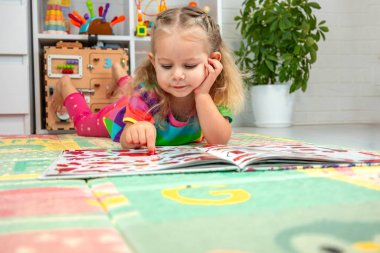 Parlak elbiseli küçük bir kız evde renkli bir paspasın üzerinde oyun oynuyor ve öğreniyor.