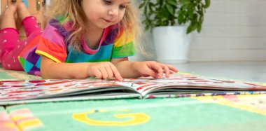 Parlak elbiseli küçük bir kız evde renkli bir paspasın üzerinde oyun oynuyor ve öğreniyor.