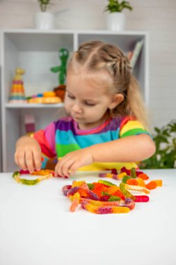 Gökkuşağı tişörtlü mutlu küçük kız renkli şekerlerle veya vitaminlerle oynuyor ve yiyor.