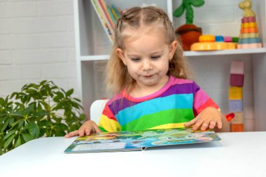 Parlak tişörtlü sevimli küçük kız kitap okuyor ve sayfayı işaret ediyor.