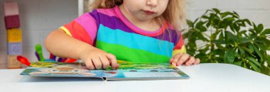 Parlak tişörtlü sevimli küçük kız kitap okuyor ve sayfayı işaret ediyor.