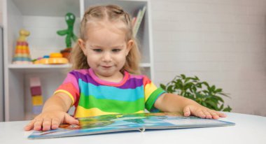 Parlak tişörtlü sevimli küçük kız kitap okuyor ve sayfayı işaret ediyor.