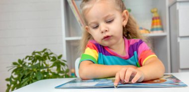 Parlak tişörtlü sevimli küçük kız kitap okuyor ve sayfayı işaret ediyor.