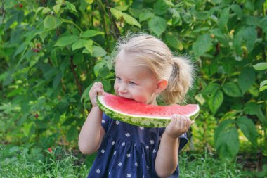 Mutlu küçük kız yeşil bir bahçenin arka planında sulu bir karpuz yiyor..