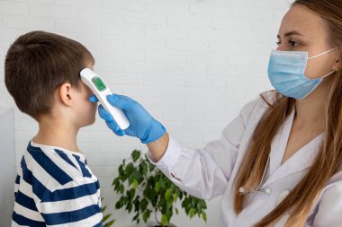 Yüz maskeli kadın doktor, çocuğun ateşini kızılötesi termometreyle ölçüyor..