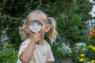 Çiçek açan bir yaz bahçesinde büyüteçle petunyalara bakan küçük bir kız..