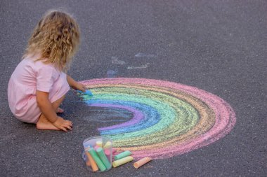 Küçük kız yazın asfalta renkli tebeşirle parlak bir gökkuşağı çiziyor.