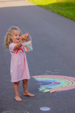 Küçük kız yazın asfalta renkli tebeşirle parlak bir gökkuşağı çiziyor.