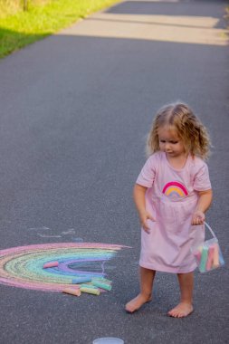 Küçük kız yazın asfalta renkli tebeşirle parlak bir gökkuşağı çiziyor.